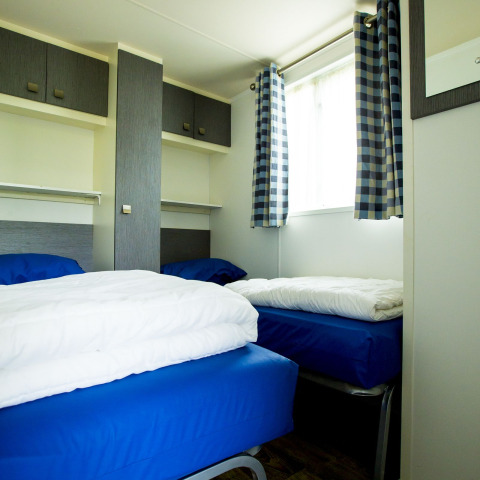 To enkeltsenge med blåt sengetøj på et værelse med skab og ternede gardiner i Delta Lodge, Holiday Park Duinhoeve.