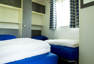 Twin beds with blue bedding in a compact room with cabinets and plaid curtains in Delta Lodge, Holiday Park Duinhoeve.