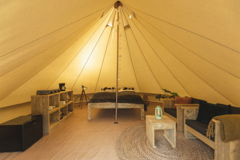 Interior de una tienda tipi con muebles de madera, cama, sofá, estantería, mini nevera y luz acogedora.