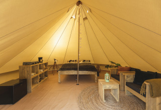 Indretning af et tipi-telt med træmøbler, seng, sofa, hylder, køleskab og hyggelig belysning.