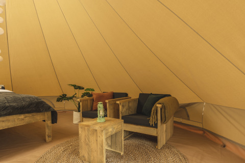 Interior of a teepee tent with wooden furniture, black cushions, a green lantern, and cozy decor.