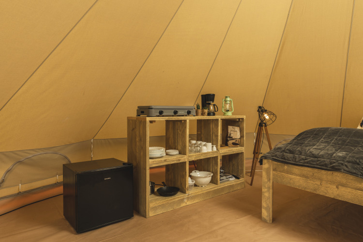 Interior of a teepee tent with a wooden shelf, mini fridge, stove, coffee maker, and cozy lighting.