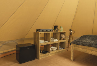 Interior of a teepee tent with a wooden shelf, mini fridge, stove, coffee maker, and cozy lighting.
