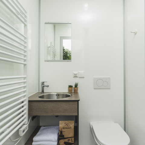 Baño moderno y compacto en una tienda tipi, con espejo, lavabo, inodoro, toallas y set de servicios visible.