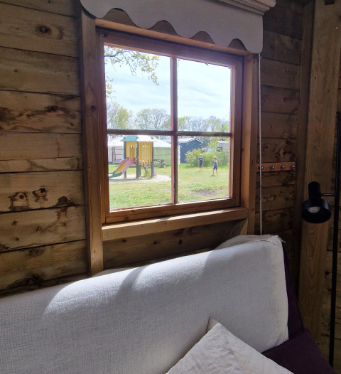 Gezicht uit het raam van een houten hut op speelplein en kinderen buiten. Writers hut bij Zomerlicht, NL.