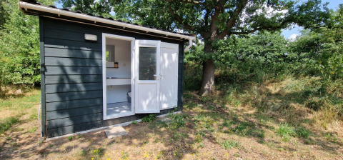 Sort træhytte med åbent dørparti ved en glampingplads, omgivet af træer og natur i solskin.
