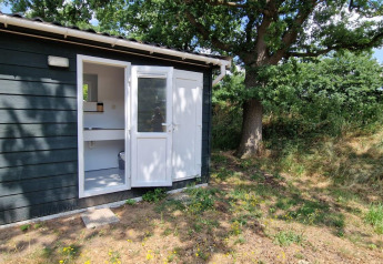 Sort træhytte med åbent dørparti ved en glampingplads, omgivet af træer og natur i solskin.