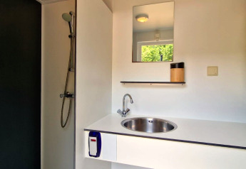 Salle de bain moderne en glamping avec douche, lavabo, miroir et étagère, vue sur la verdure.