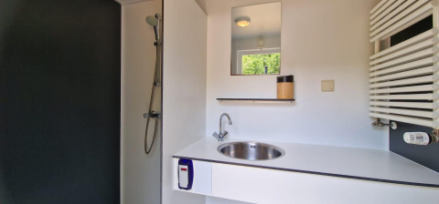 Moderne glampingbadeværelse med bruser, håndvask, spejl og hylde med udsigt til grøn natur.