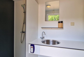 Moderne glampingbadeværelse med bruser, håndvask, spejl og hylde med udsigt til grøn natur.