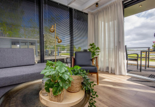 Sala moderna en una casa flotante con plantas, grandes ventanas, persianas negras y vista al agua.