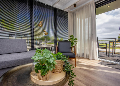 Salon moderne sur une péniche avec plantes vertes, grandes fenêtres, stores noirs et vue sur l’eau.