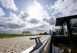 Vista de la casa flotante 'De Waterparel' en un muelle soleado de Friesland, Países Bajos, con banco y cielo.