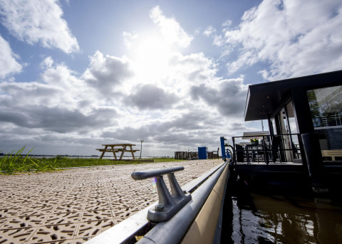Uitzicht op Houseboat 'De Waterparel' aan een zonnige steiger in Friesland, Nederland, met bankje en wolken.