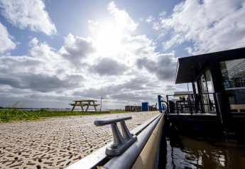 Vista de la casa flotante 'De Waterparel' en un muelle soleado de Friesland, Países Bajos, con banco y cielo.