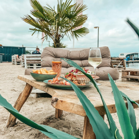 Atmosfera glamping con tavolo rustico di legno, divano morbido, palma e piatti serviti sulla sabbia.