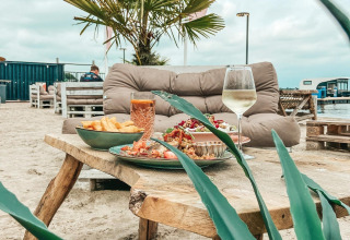 Atmosfera glamping con tavolo rustico di legno, divano morbido, palma e piatti serviti sulla sabbia.