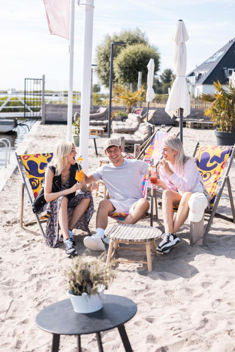 Drei junge Erwachsene entspannen auf Liegestühlen im Sand an einer Glamping-Unterkunft mit Getränken.