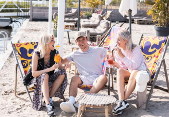 Drei junge Erwachsene entspannen auf Liegestühlen im Sand an einer Glamping-Unterkunft mit Getränken.