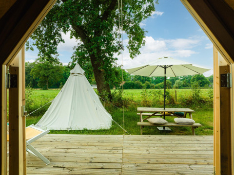 Udsigt fra en træhytte til et tipitelt, picnicbord og parasol i en grøn eng ved Holiday Park Mölke, Holland.