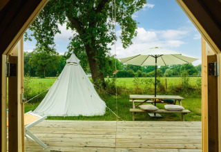 Vista desde una cabaña hacia una tienda tipi, mesa de picnic y sombrilla en un prado del Holiday Park Mölke, Países Bajos.