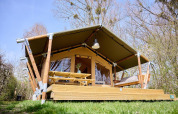 Safari telt Villatent Compact med overdækket træterrasse, picnicbord og bænk i grønne omgivelser.