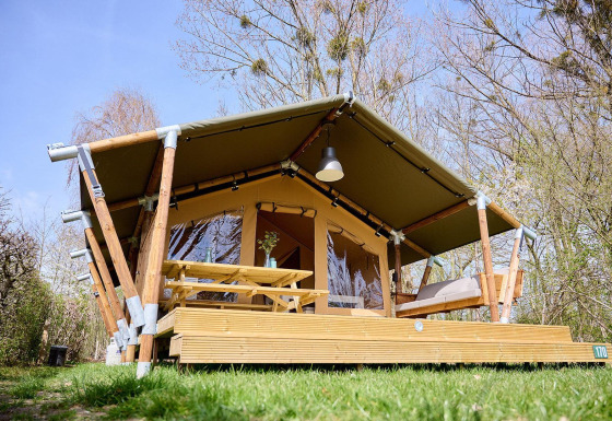 Safari tent Villatent Compact with covered wooden deck, picnic table, and bench in a green setting.