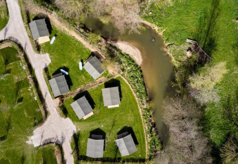 Vista aerea delle tende safari Villatent Compact lungo un fiume, circondate da prato verde e alberi sparsi.