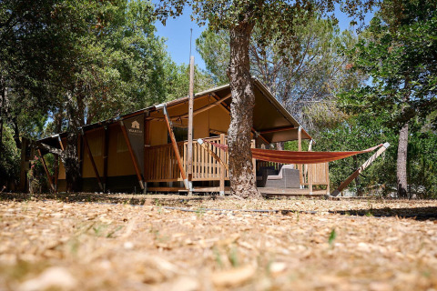 Tienda safari Villatent Nomad con terraza y hamaca en Camping Leï Suves, Francia, rodeada de árboles verdes.