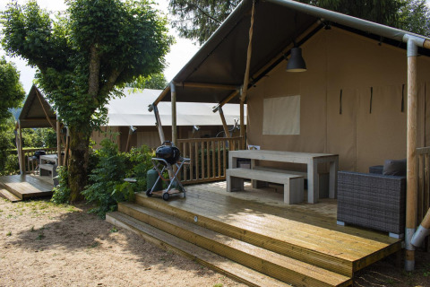 Tienda safari Villatent Nomad con terraza de madera y barbacoa en Camping Les Genêts, Francia, rodeada de naturaleza.
