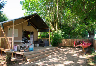 Tienda safari Villatent Nomad en Camping Les Genêts, Francia, con terraza de madera y hamaca al aire libre.