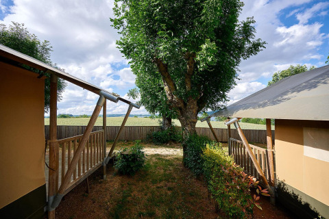 Zicht vanuit een Villatent Nomad safaritent bij Camping Les Genêts in Frankrijk met boom en houten veranda’s.