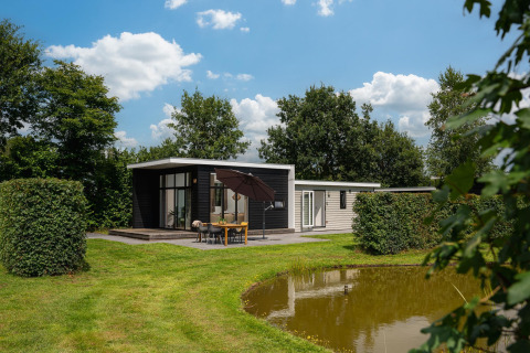 Sistemazione glamping moderna con terrazza e ombrellone vicino a uno stagno, tra alberi verdi.