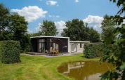 Modern glamping accommodation with terrace and umbrella by a small pond, surrounded by greenery.
