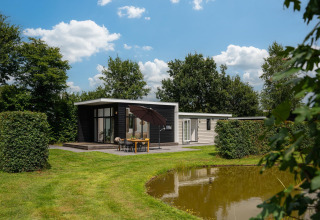 Moderne glampingaccommodatie met terras en parasol aan een vijver, omringd door groen.