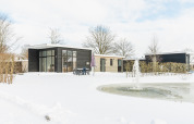 Alloggio glamping Forest Lodge in inverno, cabina moderna tra la neve con vista su stagno naturale.
