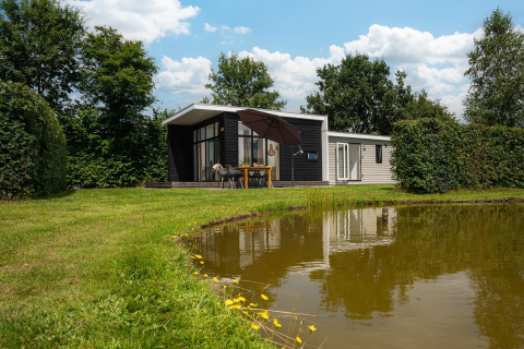 Glamping indkvartering ved en dam med moderne hytte, udendørs spisebord og grønne træer omkring.
