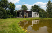 Glamping indkvartering ved en dam med moderne hytte, udendørs spisebord og grønne træer omkring.