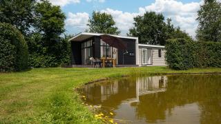 Glamping-accommodatie bij een vijver met een moderne hut, buitentafel en omringd door veel groen.