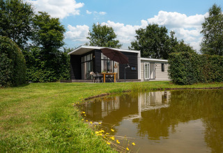 Glamping con cabina moderna vicino a uno stagno, tavolo da pranzo all’aperto e tanto verde attorno.
