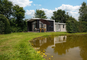 Glamping indkvartering ved en dam med moderne hytte, udendørs spisebord og grønne træer omkring.