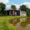 Glamping-accommodatie bij een vijver met een moderne hut, buitentafel en omringd door veel groen.