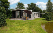 Glamping accommodation with a modern cabin, spacious terrace, outdoor dining area, umbrella, and pond view.