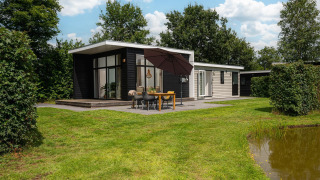 Glamping indkvartering med moderne hytte, stor terrasse, spisebord, paraply og grønne omgivelser ved en dam.