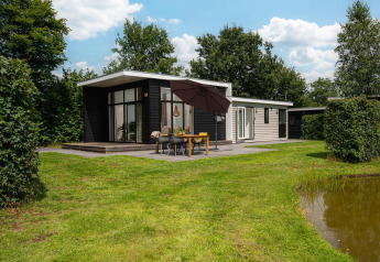 Glamping indkvartering med moderne hytte, stor terrasse, spisebord, paraply og grønne omgivelser ved en dam.