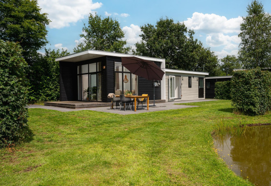 Glamping indkvartering med moderne hytte, stor terrasse, spisebord, paraply og grønne omgivelser ved en dam.