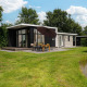 Glamping indkvartering med moderne hytte, stor terrasse, spisebord, paraply og grønne omgivelser ved en dam.