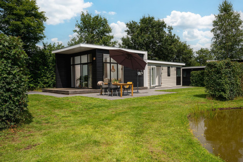 Glamping accommodation with a modern cabin, spacious terrace, outdoor dining area, umbrella, and pond view.