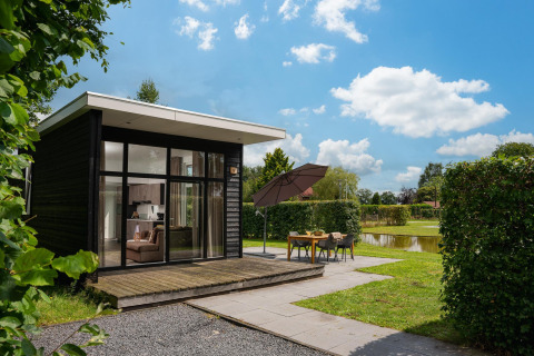 Cabaña glamping moderna con grandes ventanales, terraza y comedor exterior junto a un estanque bajo cielo azul.