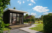 Moderne glamping-hytte med store vinduer, terrasse og udendørs spisested ved dam under blå himmel.
